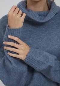 Person, der die Ärmels eines weichen, blauen Strickrollkragenpullis anpasst, zeigt eine Nahaufnahme von Händen und Oberkörper.