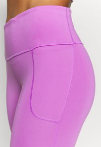 Detailní záběr osoby v levandulových legínách s vysokým pasem, v nichž jsou viditelné stehy boční kapsy.