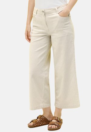 Pantalones de pana beige de pierna ancha usados con sandalias marrones con doble tirante y tachuelas, una mano en el bolsillo, torso inferior visible.
