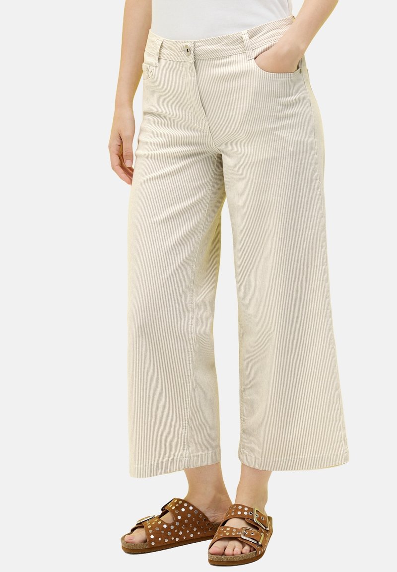 Pantalon large beige en velours côtelé porté avec des sandales marron à doubles brides cloutées, une main dans la poche, torse inférieur visible.