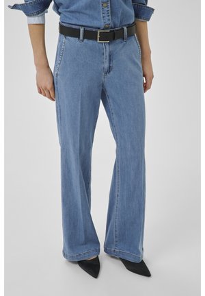 Személy világoskék, bő szárú farmernadrágban, fekete övvel és fekete hegyes orrú cipőben, laza kéztartással oldalán áll.