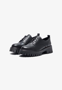 Des chaussures en cuir noir présentent un bout rond, six oeillets, et une semelle épaisse et dentelée pour plus d'adhérence. Texture lisse avec un minimum de coutures.