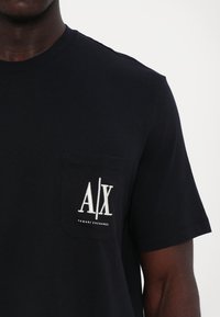 Fekete, rövid ujjú pamut póló, mellzsebében fehér "A/X" logóval és alatta "Armani Exchange" felirattal.