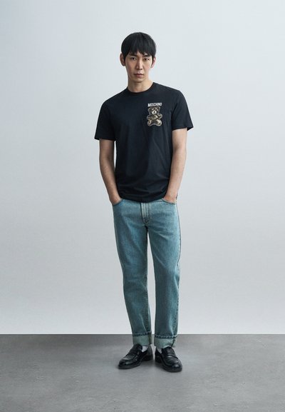 Μαύρο βαμβακερό μπλουζάκι με γραφικό αρκούδας στην αριστερή τσέπη του στήθους, σε συνδυασμό με ανοιχτόχρωμο τζιν παντελόνι και μαύρα παπούτσια.