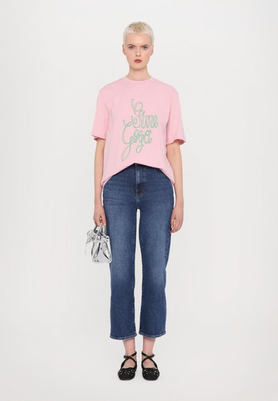 Camiseta de algodão rosa com design em verde, combinada com jeans de perna reta azuis. Os acessórios incluem uma bolsa transparente e sapatos pretos com tiras cruzadas.