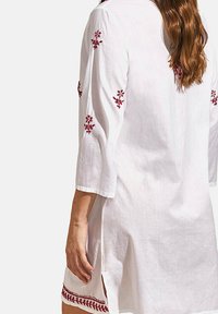 Tunique en lin blanc avec des manches trois-quarts. Présente une broderie florale rouge sur les manches et l'ourlet, ainsi que des fentes latérales pour un meilleur confort.