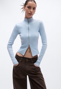 Γαλάζιο μπλουζάκι με μακριά μανίκια, ψηλό λαιμό και φερμουάρ στο μπροστινό μέρος· διαθέτει κοντό μήκος και ριμπ υφή, συνδυασμένο με καφέ παντελόνια με φαρδιά πόδια.