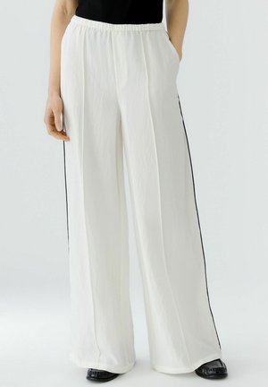 Weiße weit geschnittene Hose mit elastischem Bund und schwarzen Seitenstreifen, getragen mit schwarzen Schuhen und einem schwarzen Oberteil.