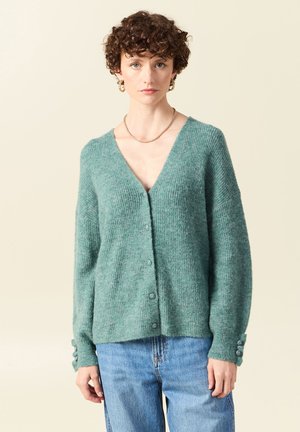 Cardigan verde texturizzato con scollo a V, bottoni frontali e dettagli con tre bottoni sulle maniche. Indossato sopra jeans azzurri chiari.