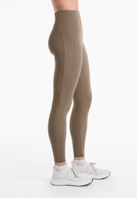 Magas derekú barna leggings, bordázott textúrájú panelek a szélek mentén, fehér sportcipőkkel és világosszürke zoknikkal párosítva.