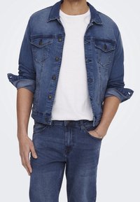 Donkere blauwe spijkerjas, met een klassieke kraag, knoopsluiting aan de voorkant en twee borstzakken. Gedragen over een wit T-shirt en gecombineerd met bijpassende jeans.