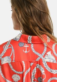Maglietta rossa con motivi nautici blu navy, inclusi corde, conchiglie e un'ancora. Presenta un colletto e una piega sul retro per un design aggiuntivo.