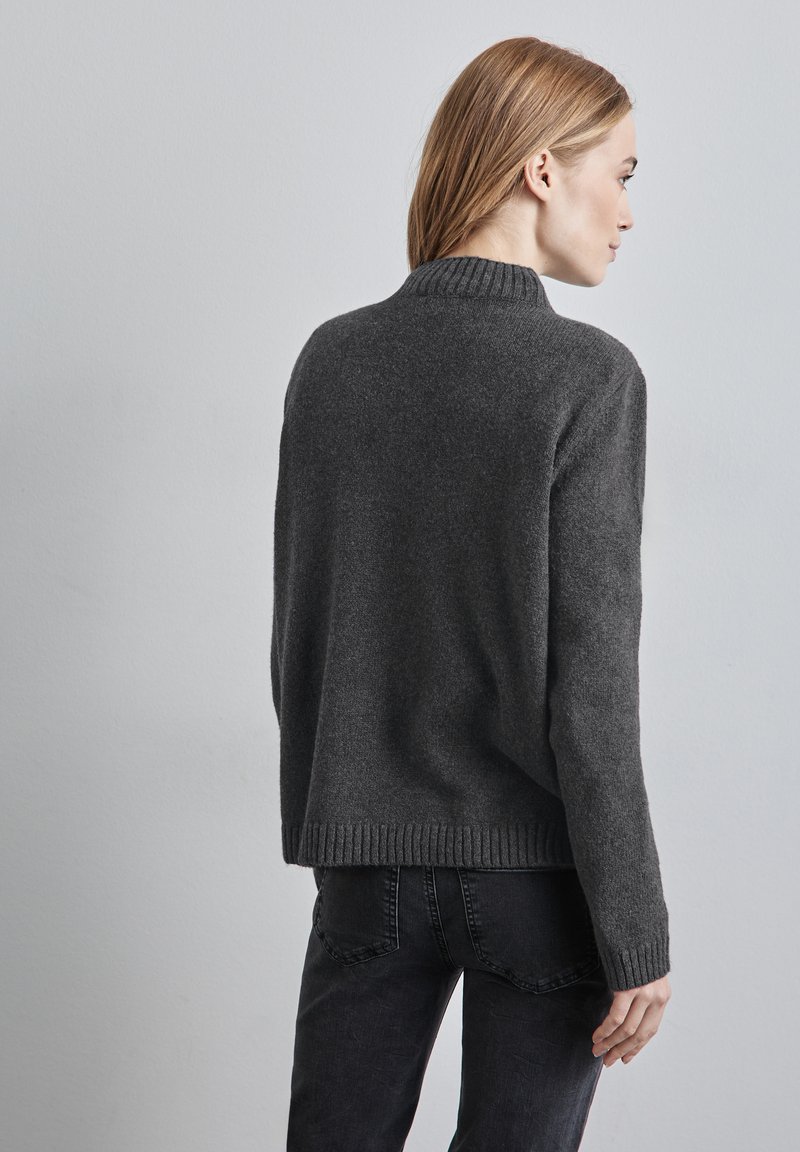 Grå stickad tröja med hög ribbad krage, sänkt axelsöm och ribbad nederkant, tillsammans med mörka jeans. Slät textur, avslappnad passform.