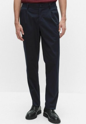 Man i marinblå skräddarsydda byxor med sidofickor och svarta snörskor, stående mot en enkel ljus bakgrund.