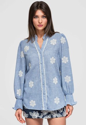 Femme portant un chemisier bleu à manches longues avec des motifs de fleurs blanches et une bordure en dentelle, associé à une jupe noire brodée de fleurs.