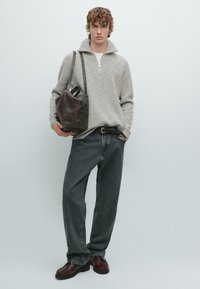 Giovane uomo che indossa un maglione grigio a coste, jeans scuri, scarpe in pelle marrone, mentre porta a tracolla una borsa tote in pelle marrone scuro.