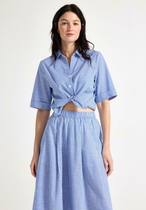 Ung kvinde med mørkt hår iført en blå og hvidternet kortærmet skjorte, der er knyttet ved taljen, og matchende høj-taljede nederdel.