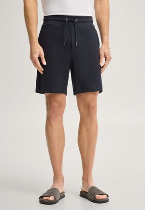 Mann trägt schwarze, gestrickte Shorts mit Kordelzug und graue offene Zehen-Pantoffeln und steht vor einem schlichten Hintergrund.