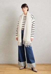 Écharpe crème avec des bords frangés, portée sur un pull rayé bleu marine et blanc. Jean en denim à jambe large avec des poignets gris contrastants. Chaussures gris clair.