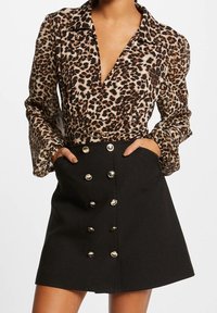Bluse med leopardmønster, lange ærmer og dyb V-udskæring, parret med en sort nederdel med guldknapdetaljer og sidelommer.
