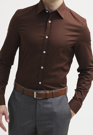 Homme portant une chemise ajustée marron foncé à boutons, un pantalon gris et une ceinture en cuir marron avec une boucle argentée.