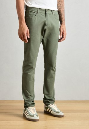 Grüne Baumwollmischungshose mit schlankem Schnitt, ausgestattet mit Vordertaschen und einem Bund mit Gürtelschlaufen. Kombiniert mit grau-grünen Sneakers.