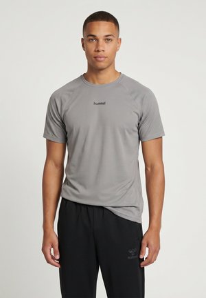 Ung mand står iført en grå kortærmet sportst-shirt og sorte træningsbukser mod en ensfarvet lys baggrund.