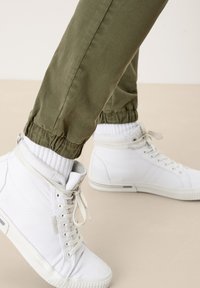Persoon met witte hoge sneakers, geribbelde witte sokken en olijfgroen joggingbroek die op een beige vloer staat.