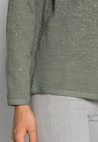 Oliivinvihreä neulepaita, jossa on tekstuuri, pitkät hihat ja pyöreä kaula-aukko, joka on yhdistetty vaaleanharmaisiin housuihin.