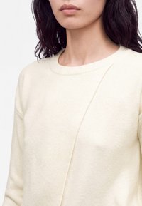 Femme aux cheveux foncés portant un pull crème avec un détail de couture diagonale à l'avant, visible des épaules jusqu'au milieu du torse.