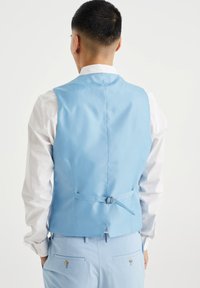 Jasnoniebieska kamizelka z tkaniny o satynowym wykończeniu, z klamrą z tyłu i gładkim, dopasowanym krojem, noszona na białej koszuli.