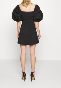 Czarna krótka sukienka z bufkami, kwadratowym dekoltem z tyłu, dopasowanym gorsetem, rozkloszowaną spódnicą i subtelną teksturą. Noszona z czarnymi szpilkami.