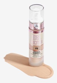Base de maquillaje de larga duración Revolution IRL Filter en el tono F9, presentada en un frasco de plástico transparente con acentos en rosa, junto a una muestra de la base líquida.