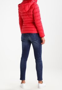 Czerwona kurtka puchowa z kapturem, zapinana na zamek, z poziomymi przeszyciami, połączona z ciemnoniebieskimi, dopasowanymi d�żinsami i białymi sneakersami.