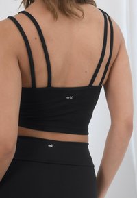 Top corto nero con multiple cinghie sottili sul retro, caratterizzato da un logo. Il tessuto sembra liscio e elastico, abbinato a pantaloni neri coordinati.