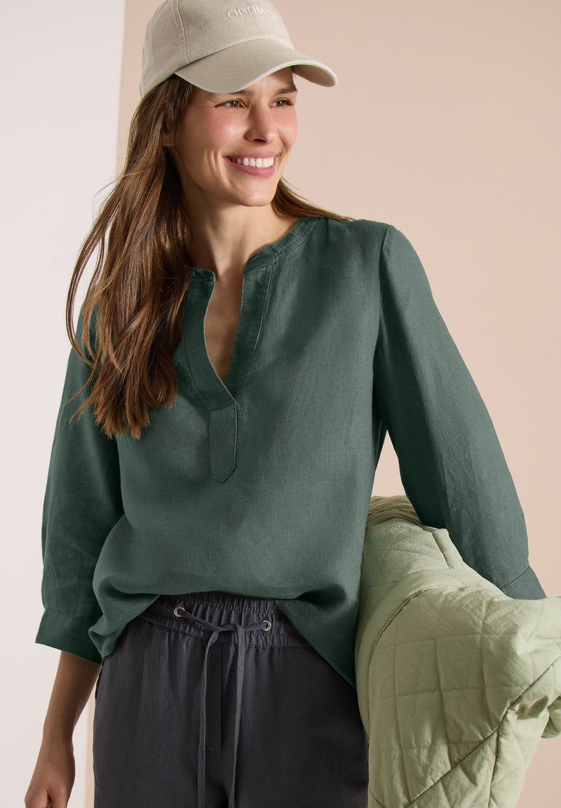 Femme souriante portant une casquette beige, un chemisier vert foncé à col en V et un pantalon foncé à cordon, tenant une veste matelassée vert clair sur le bras.
