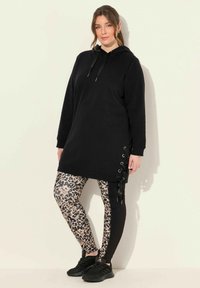 Černé šaty mikinového stylu s postranními šňůrkami, kombinované s černými legínami s leopardím potiskem. Modelka má na sobě černé tenisky.