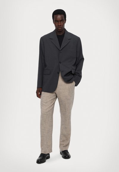 Hallikas üle mõõdu suur blazer ühe nööbiga, ühe rinnataskuga; koos heleda beeži mustriga pükstega ja mustade kingadega.