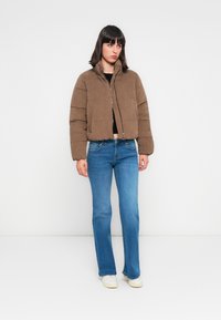 Veste matelassée marron avec un col haut, fermeture éclair et tissu texturé. Associée à un jean clair évasé et des baskets blanches.