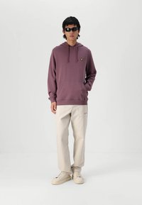 Sudadera burdeos con capucha y bolsillo delantero, combinada con pantalones crema y zapatillas blancas. Los accesorios incluyen gafas de sol oscuras. Diseño minimalista.