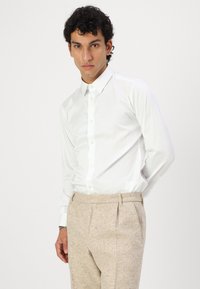 White long-sleeve shirt with button-down collar, paired with beige textured trousers. Shirt features a smooth, fitted design.