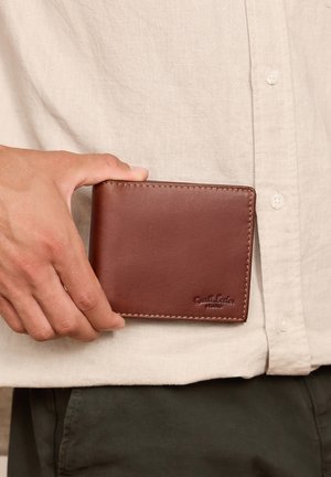 Main tenant un portefeuille bifold en cuir marron devant une chemise beige boutonnée et un pantalon foncé.