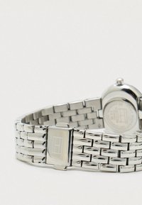 Sudraba metāla pulksteņa aproce ar locīšanu, kurā ir redzams taisnstūra logo uz locīšanas mehānisma, uz vienkārša fona.