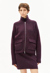 Frau mit kurzbraunem Haar, die eine gesprenkelte lila Zip-Jacke mit großen Fronttaschen und einem passenden Rock vor einem weißen Hintergrund trägt.