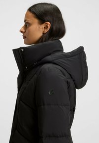 Svart puffer-jacka med hög krage, fast huva och sidodragkedja. Texturerat tyg med en logotyp på vänster ärm.