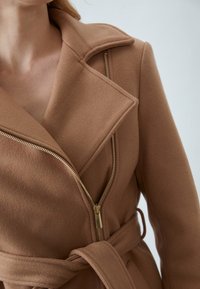 Manteau en mélange de laine marron avec un col cranté, doté d'une fermeture éclair dorée visible et d'une ceinture en tissu assorti, texture lisse et coupe cintrée.