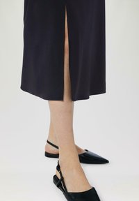 Chaussons noirs à bout pointu avec un design de brides, associés à une robe sombre longueur cheville avec une fente sur le côté. Texture lisse, silhouette élégante.