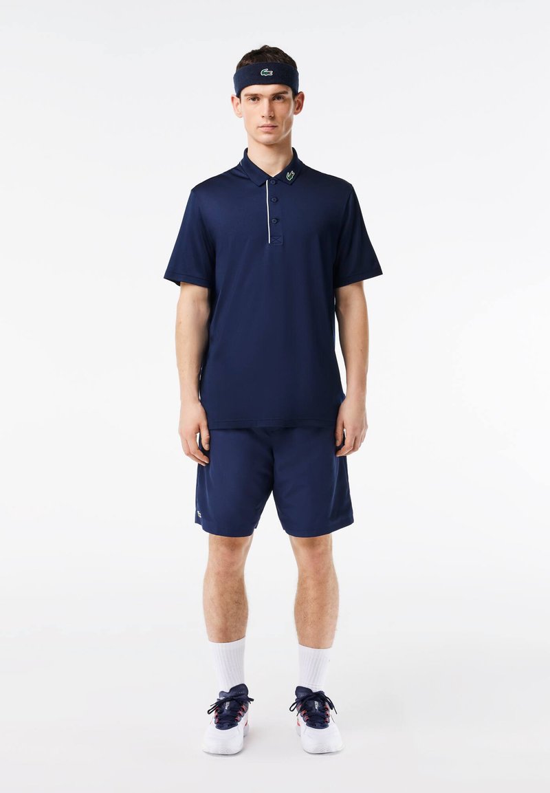 Ναυτικό πόλο μπλουζάκι με τρία κουμπιά και αντίθετο γιακά, συνδυασμένο με κοντές σορτς. Περιλαμβάνει ένα κορδέλα για το κεφάλι με λογότυπο. Λευκές κάλτσες και αθλητικά παπούτσια.