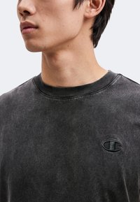 Gros plan sur une personne portant un t-shirt noir délavé à col rond avec un petit logo Champion brodé sur le côté gauche de la poitrine.
