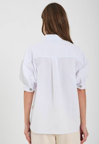 Camicia bianca con maniche a sbuffo, polsini abbottonati e dettaglio della yoke sul retro. Realizzata in tessuto morbido, con orlo curvo.